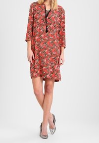 Punainen mekko mustilla ruusukuvioilla, 3/4-hihoilla, V-pääntiellä, jossa on nyöritys, polven pituinen, yhdistettynä hopeisiin korkokenkiin.