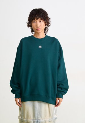 Person med kort krøllete hår iført en tealfarget Adidas-genser og lys denimskjørt foran en ensfarget hvit bakgrunn.