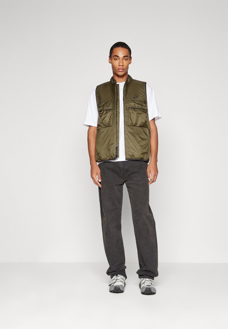 Gilet matelassé vert olive avec fermeture éclair à l'avant, deux poches poitrine et un petit logo, porté sur un T-shirt blanc et un pantalon noir, associé à des baskets grises.