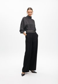 Schwarz-weiße Bluse mit Polka-Dots und hohem Kragen, kombiniert mit maßgeschneiderten schwarzen weitgeschnittenen Hosen und schwarzen spitzen High Heels.