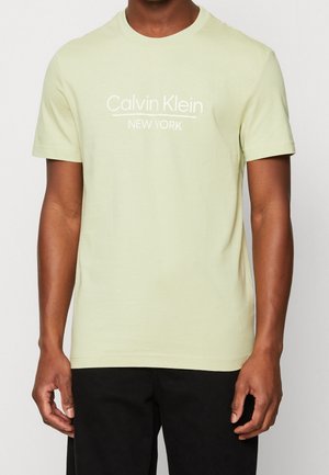 Mann trägt ein hellgrünes T-Shirt mit kurzen Ärmeln mit weißem „Calvin Klein New York“-Schriftzug, kombiniert mit schwarzen Hosen, steht vor weißem Hintergrund.