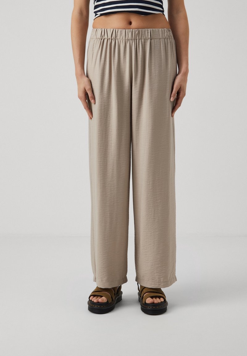 Femme portant un pantalon beige ample, des sandales noires à plateforme et un haut court rayé noir et blanc, debout devant un fond neutre.