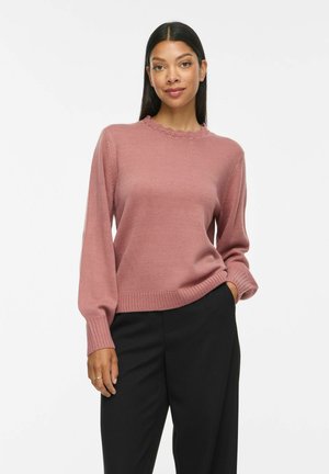 Femme debout avec une main dans la poche, portant un pull en maille rose poudré avec un col festonné et un pantalon noir.