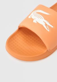 Πορτοκαλί λαστιχένιο σανδάλι slide με ανάγλυφο πέλμα και φαρδιά λουρά με λευκό λογότυπο κροκοδείλου. Τετράγωνο σχέδιο στη σόλα.