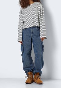 Person, der ein langärmliges gestreiftes Shirt in Weiß und Schwarz, loose blaue Cargo-Jeans und braune Schnürstiefel trägt, steht vor einem schlichten Hintergrund.