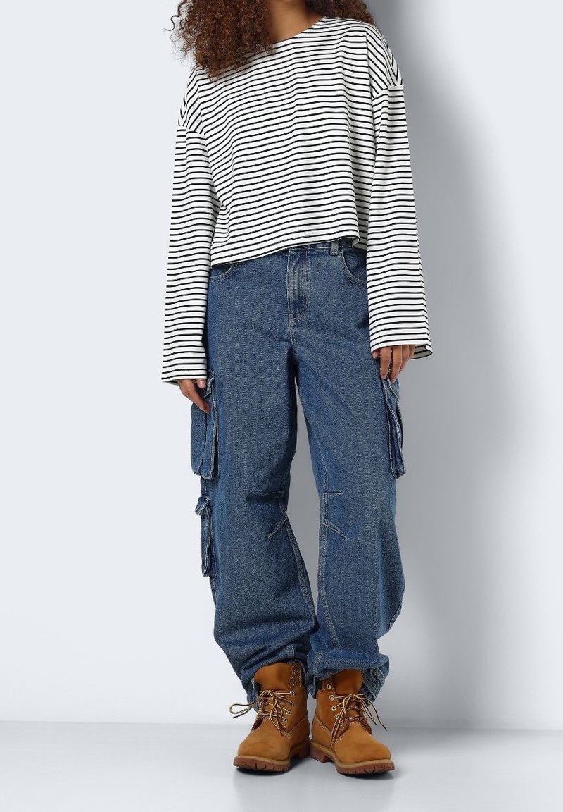 Person, der ein langärmliges gestreiftes Shirt in Weiß und Schwarz, loose blaue Cargo-Jeans und braune Schnürstiefel trägt, steht vor einem schlichten Hintergrund.