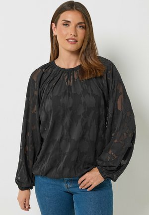 Frau mit schulterlangem braunem Haar, trägt eine durchsichtige schwarze Bluse mit Blumenmuster und blaue Jeans, leicht lächelnd, Hand auf der Hüfte.