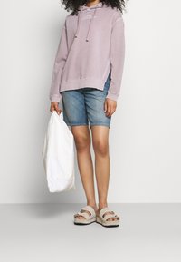 Sudadera lila con logo, pantalones cortos de mezclilla azules, sandalias de plataforma crema con hebilla y un bolso tote blanco. Atuendo casual.