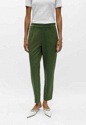 Femme portant un pantalon taille haute vert olive ajusté, un top blanc à manches courtes, des chaussures slingback blanches à bout pointu et un bracelet argenté au poignet gauche.