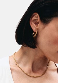 Pendientes de aro dorados con superficie texturizada, combinados con un delicado collar de cadena dorada con un diseño retorcido, usados sobre un fondo neutro.