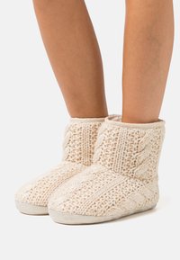 Gehaakte, crème-kleurige slippers met een kabelpatroon, zachte textuur en gevoerde zolen. De korte hoogte bedekt de enkel comfortabel.
