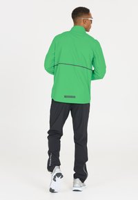 Veste légère verte avec un col haut et une bande réfléchissante ; associée à un pantalon noir et des chaussures de sport.