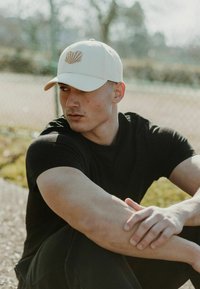Beige sapka, elülső részén kagyló emblémával, egy fekete pólót viselő személynél. A háttérben elmosódott zöld növényzet és egy kerítés látható.