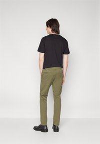 T-shirt nera in cotone, pantaloni slim-fit verde oliva e scarpe nere, visti da dietro su uno sfondo bianco semplice.