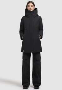 Eine Frau steht mit den Händen in den Taschen, trägt einen schwarzen winterlichen Kapuzenmantel, schwarze Cargo-Hosen und schwarze Schuhe vor einem einfarbigen hellen Hintergrund.