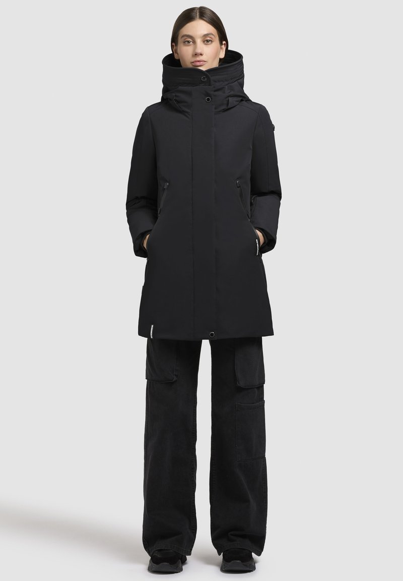 Eine Frau steht mit den Händen in den Taschen, trägt einen schwarzen winterlichen Kapuzenmantel, schwarze Cargo-Hosen und schwarze Schuhe vor einem einfarbigen hellen Hintergrund.