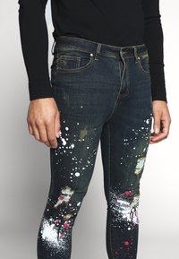 Jeans de mezclilla oscura desgastada con detalle de salpicaduras de pintura en blanco y rojo, con roturas y un ajuste ajustado. Incluye bolsillos clásicos y cierre de botón.