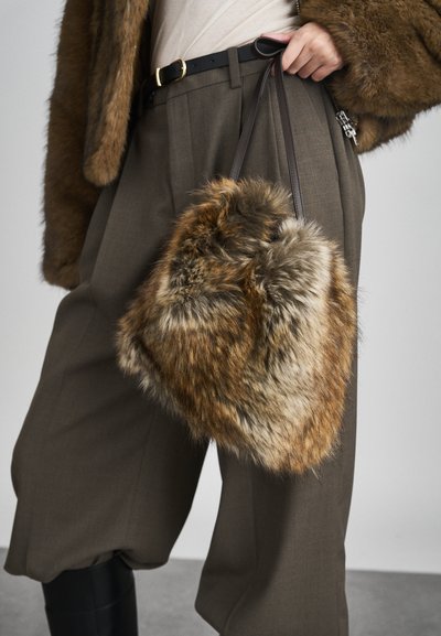 Brūnas mākslīgās ādas rokassoma ar mīkstu tekstūru, kurai ir tumša roktura. Valkāta ar pielāgotām pelēkām biksēm un gaišu augšu. Minimālistisks dizains.