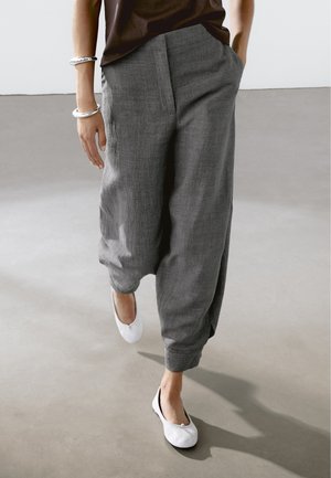 Person walking wearing loose gray pants, white ballet flats, dark top, and silver bracelets, with one hand in pocket.