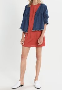 Džínová krátká bunda s třepeným lemem, přes jednobarevné červené shiftové šaty. Bílé nazouvací boty. Modelka stojí na bílém pozadí.