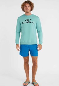 Mätna zelená mikina s čiernym logom "O'Neill", doplnená modrými šortkami. Neformálny vzhľad doplnený svetlými sandálmi.