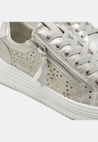 Metallic silver sneaker with floral perforations, lace-up front, and side zipper. Thick white sole with horizontal grooves and texture.