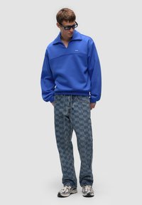 Pullover in fleece blu con colletto ripiegato, abbinato a pantaloni in denim chiaro con un motivo a logo ripetuto. Il modello indossa occhiali da sole riflettenti.