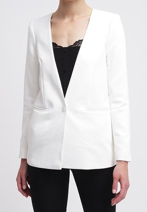 Femme portant un blazer blanc ajusté par-dessus un top noir bordé de dentelle et un pantalon noir, debout devant un fond uni.