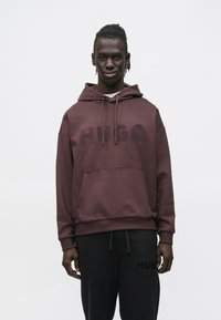 Felpa con cappuccio color marrone scuro con grande logo "HUGO" nero, cappuccio con coulisse, tasca frontale e vestibilità comoda. Abbinata a pantaloni della tuta neri con logo.