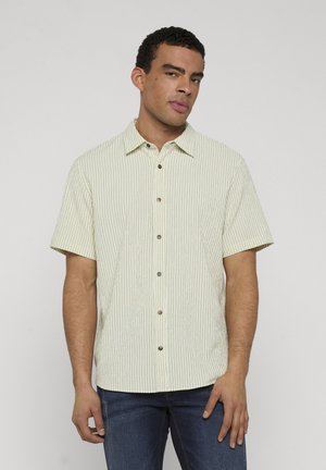 Mężczyzna w jasnobeżowej, krótki rękaw, koszuli w paski na guziki i ciemnoniebieskich dżinsach, stojący na jednolitym białym tle.