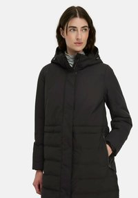 Frau in einer schwarzen, kapuzenbewehrten Steppjacke mit seitlichem Reißverschlusstaschen, die gegen einen schlichten weißen Hintergrund zur Seite blickt.