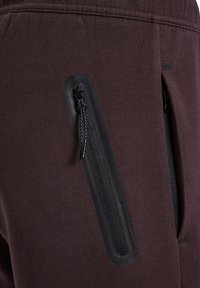 Mørkebrune joggers med to lodrette lynlåslommer med sort hardware. Blød tekstur, elastisk taljebånd og minimal design.