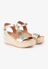 Sandalias de cuña metálicas doradas con un acabado texturizado, tira de tobillo ajustable y una suela trenzada al estilo espadrille. Base de plataforma plana.