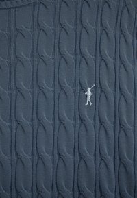 Blauer Strickstoff mit wiederholtem Zopfmuster und einer kleinen weißen, bestickten Figur einer Person, die eine Golfschläger trägt.