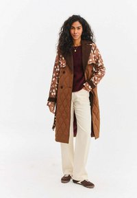 Brązowy pikowany trencz w cętkowany wzór, z dwurzędowymi guzikami i kontrastującym brązowym kołnierzem. W zestawieniu z kremowymi spodniami i brązowymi butami.