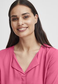 Pink blouse with a soft, lightweight texture, featuring a V-neckline and side seam detail, displaying a relaxed fit and smooth fabric.