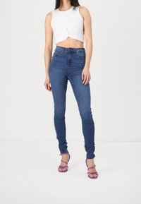 Bílý zkrácený top s uzlem vpředu, vysoké modré skinny džíny a fialové páskové podpatky. Hladká látka, těsně přiléhající střih.