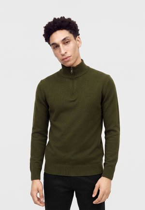 Jeune homme aux cheveux bouclés portant un pull vert olive à fermeture éclair quart, et un pantalon noir, debout devant un fond clair uni.