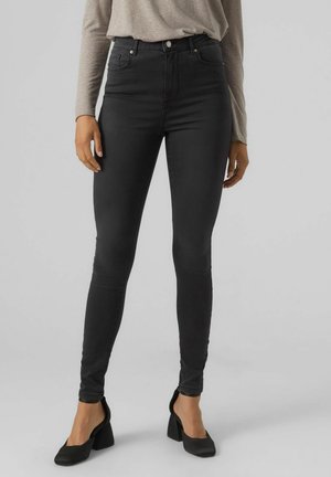 Jeans negros de tiro alto con ajuste ceñido, confeccionados en denim elástico. Cuentan con bolsillos frontales y una cremallera con botón. Se llevan con zapatos de tacón negros.