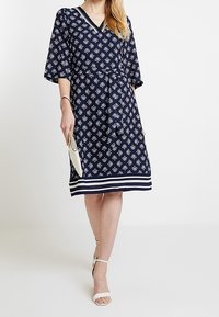 Marinblå ombuktsklänning med vitt blommönster, V-ringning, trekvartsärmar och randig nederkant. Matchad med vita klackar och en kuvertväska.