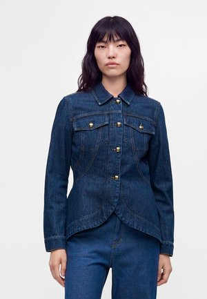 Frau mit langen schwarzen Haaren, die eine dunkelblaue Jeansjacke mit goldenen Knöpfen und passende Jeanshosen vor einem schlichten weißen Hintergrund trägt.