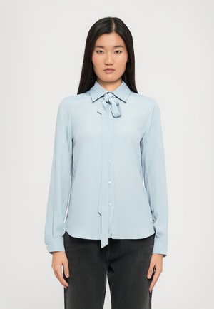 Blouse bleu clair à manches longues avec un col et un détail de nœud, fabriquée dans un tissu lisse. Boutonnage sur le devant, ourlet incurvé et manches à poignets.