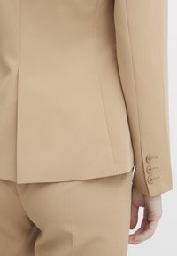 Beige op maat gemaakte blazer met een getailleerde silhoeet; heeft een ventiale rug, knopen met drie knopen en een gladde stoftextuur.