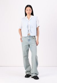 Lyseblå quiltet vest med båndknute, båret over en hvit skjorte; kombinert med lyseblå flare-jeans og svarte kraftige sko.