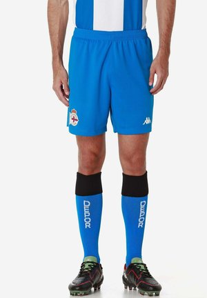 Homme portant un short de football Deportivo bleu, des chaussettes bleu assorties avec des dessus noirs et des crampons noirs avec des touches rouges, debout sur fond blanc.