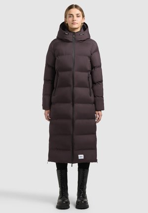 Femme portant un long manteau matelassé marron foncé avec une capuche, des leggings noirs et des bottes noires à lacets, debout devant un fond clair uni.