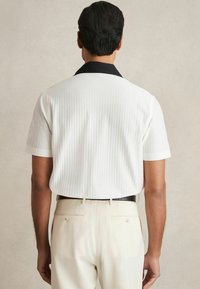 Cămașă cu mânecă scurtă, de culoare albă cu guler negru, având o textură ribbed verticală. Asortată cu pantaloni bej, prezentând vedere din spate.