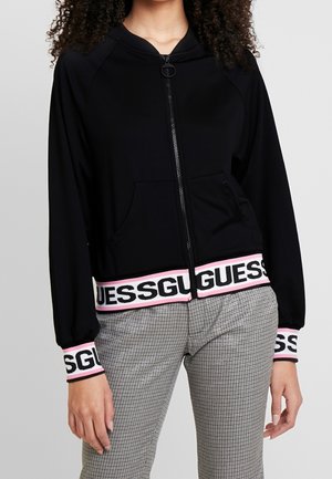 Kobieta ubrana w czarną bluzę z kapturem zapinaną na zamek, z logo "GUESS" na mankietach i w pasie, w połączeniu z szarymi spodniami w kratę, stojąca na szarym tle.