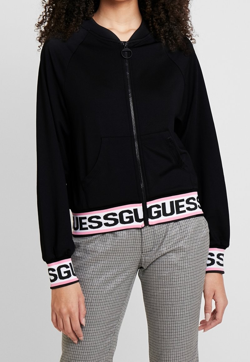 Femme portant un sweat à capuche noir zippé avec le logo "GUESS" sur les poignets et la ceinture, associé à un pantalon gris à carreaux, debout devant un fond gris.