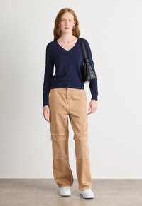 Maglione a v in navy abbinato a pantaloni cargo larghi color sabbia con cuciture sulle ginocchia. La modella porta una piccola borsa nera e indossa scarpe da ginnastica bianche.